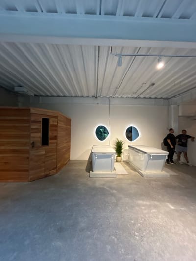 The image shows the interior of a spa facility located in Pozos, Costa Rica (9.952, -84.196). Two white, rectangular, tubs are placed in the center of the room, with a plant in between them. The room has a white, tiled ceiling with track lighting. There is a wooden sauna to the left, and two oval mirrors on the far wall, both reflecting greenery. Two men are standing in the background, to the right of the tubs. The floor is concrete, and the room is illuminated by overhead lighting.