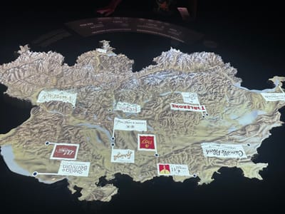 The image shows a topographical map of Switzerland with several chocolate companies overlaid on the map. The city of Kilchberg (ZH), Switzerland is not explicitly identified, but the Lindt & Sprüngli logo is positioned in the general vicinity. The map features logos of various Swiss chocolate brands, including Toblerone, Cailler, Frey, Camille Bloch, Favarger, Lindt & Sprüngli, Stella Bernrain, Maestrani, Feichlin, Sprüngli, and Gys. There are also smaller logos for Sunray and Chocolats Favarger. Above the map, a panel displays statistics about the Swiss chocolate industry, including data on employees, chocolate production, exports, and sales. Two people are visible at the top of the image, interacting with the display. The lighting highlights the relief of the map.