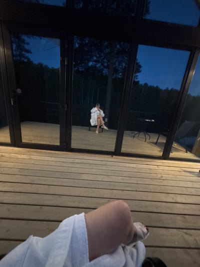 The image depicts an outdoor evening scene in Mishkino, Russia, viewed from a low perspective on a wooden deck. In the immediate foreground, the lower portion of a person's body is visible, clad in a white terry cloth robe, with a foot exhibiting red painted toenails. The foreground surface is a light brown wooden deck composed of horizontal planks. The midground features a modern structure with multiple dark-framed, tall glass panels. These panels reflect a dark blue twilight sky and dark, silhouetted trees. Through the glass, a second individual is visible indoors, seated on what appears to be a light-colored bench or chair. This individual is also wearing a white terry cloth robe. Further to the right and reflected within the interior space, a dark, spindly-legged table with a few small, indistinct objects on its surface is present. The overall lighting is dim, indicative of dusk or early night, with the interior space slightly more illuminated than the exterior.