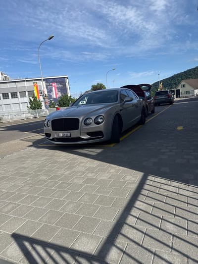 The image depicts an outdoor scene in Lausen, Canton of Basel-Landschaft, Switzerland, under a clear blue sky with scattered white clouds and bright sunlight, casting distinct shadows.
In the foreground, a silver/grey Bentley Flying Spur sedan (Swiss license plate BL-228 903, indicating Basel-Landschaft) is parked at an angle on grey interlocking paver stones. Yellow lines delineate parking spaces on the pavement. Behind the Bentley, a dark-colored sedan with its trunk open is visible. Further back, a dark grey Audi Q5 SUV (Swiss license plate SO 92853, indicating Solothurn) is also parked.
To the left, a multi-story modern building with a grey facade and horizontal windows is present. On this building, a large banner displays partial text "We m... your watches t..." and a prominent Swiss flag (red with white cross). Adjacent to the Swiss flag, a banner in yellow, red, and black, featuring an emblem (likely representing Basel-Landschaft) and a political poster with a man's face and the word "JA" (yes) are visible. In front of this building, a light-colored metal fence runs horizontally. A street light with a curved arm is positioned to the left.
To the right, a smaller, light-colored house with a brown roof is situated against a densely forested green hill, which dominates the background. Several trees are also present around the buildings. Strong, parallel shadows from a nearby railing or grate are cast across the foreground pavement from the left side of the frame. No individuals are visible in the scene.