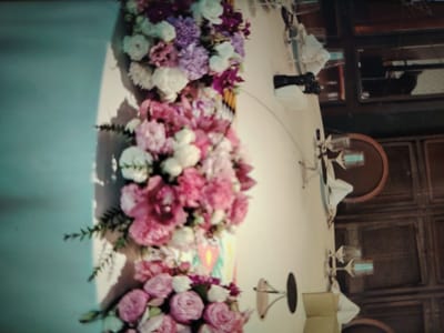 A dimly lit, indoor scene features a long table adorned with elaborate floral arrangements. The flowers are a mix of pink, white, and purple, with lush greenery. The table is set with white napkins, glasses, and what appear to be candle holders. In the background, reflections in a dark surface suggest mirrors or windows showing other parts of the establishment, possibly with seating or decorative elements. The overall ambiance is elegant and possibly festive.