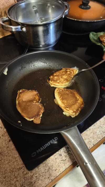The image shows a cooking scene, likely in a home kitchen in Ermolino, Russia. The primary focus is a black frying pan on a black induction stovetop, where three golden-brown pancakes are being cooked. One pancake is being flipped with a metal spatula, indicating the cooking process is in progress. The pan contains oil, and some batter has dripped onto the stovetop.
In the background, a large stainless steel pot with a glass lid sits on another burner of the stovetop. To the right of the pot, a dark orange pot is visible. A green decorative plate with a stack of pancakes can be seen on the right side of the image, suggesting these might be additional servings or preparations.
The stovetop has the brand name "Maunfeld" clearly visible. The kitchen counter appears to be made of speckled material. The overall impression is of a casual, domestic activity, possibly breakfast or brunch preparation. There are no people visible in the frame. The lighting suggests it is daytime, though no specific weather cues are apparent. The image is framed from a high-angle perspective, looking down into the pan.