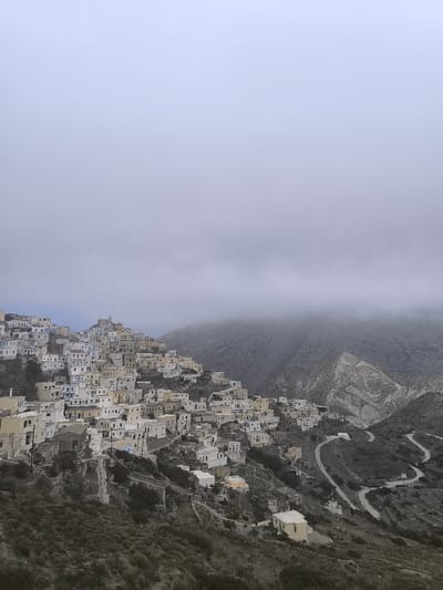 The image depicts a panoramic view of the village of Olympos, located on the island of Karpathos, Greece. The village is built on a steep hillside, with dense clusters of white and light-colored houses cascading down the slopes. Many of the houses have flat roofs and appear to be made of stone or stucco. Narrow, winding paths and staircases are visible between the buildings, indicative of a traditional Cycladic or Dodecanese architectural style.

In the distance, the landscape is dominated by rugged, mountainous terrain. A layer of thick, grey clouds hovers low over the mountains, partially obscuring the peaks and lending a dramatic, somewhat somber atmosphere to the scene. A winding road with multiple switchbacks can be seen descending the hillside on the right side of the image, leading away from the village. A small, white domed church is situated near this road.

The vegetation in the foreground and lower slopes appears sparse, consisting of low-lying shrubs and scrub. The sky is overcast and light grey, contributing to the muted color palette of the overall image. There are no people visible in the frame. The overall impression is one of a remote, traditional village nestled within a dramatic and somewhat austere natural landscape.