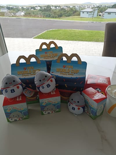 This image captures a close-up view of a table with several McDonald's Happy Meal boxes and plush toys, set against a backdrop of a scenic view of Doirí Beaga, Ireland. The McDonald's Happy Meal boxes are designed with a "FIFA World Cup" theme, featuring a blue sky and a soccer field graphic. Three of the boxes are upright, showcasing the McDonald's arches logo and the FIFA World Cup branding.

In front of the boxes, four plush toys are arranged. These toys are of a gray, shark-like character wearing soccer jerseys with the number "8" on them. Two of the plush toys are positioned in front of the boxes, while the other two are slightly behind.

To the right of the Happy Meal boxes, a red box with "BIG MAC" branding is visible. Further to the right, a portion of a McDonald's drink cup with a yellow and white straw can be seen. Some of the Happy Meal boxes have red and white checkered baskets inside.

The background of the image reveals the outdoor environment of Doirí Beaga, Ireland. Houses with light blue roofs and walls are scattered across a green landscape, with rolling hills in the distance under a cloudy sky. The foreground of the image shows a white marble-patterned table where the items are placed. The overall scene suggests a collection of McDonald's promotional items displayed on a table, with the picturesque Irish landscape serving as the backdrop.