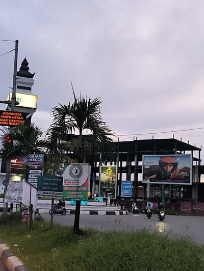 The image captures an evening street scene at an intersection in Lamprehlam Teungoh, Indonesia, under an overcast sky. In the foreground, a grassy roadside and median separate the viewer from the road. Several motorcyclists, wearing helmets, are visible on the street, some turning right around a small circular island.
To the left, a tall structure resembling a mosque minaret rises against the sky, alongside a prominent digital traffic light. The light is red, and a digital sign warns, "MENEROBOS LAMPU MERAH TIDAK MEMBUAT ANDA HEBAT!" (Running a red light does not make you great!). Below, various static signs advertise "GRAHA MECCA Percetakan" (Mecca Printing) and "BSI PRIOR."
In the mid-ground, numerous large billboards and smaller signs create a busy commercial environment. Lush palm trees are interspersed among the signs. One large green banner advertises "INGIN JAYA QURBAN" for "LAMBARO, ACEH BESAR," with prices starting from "HARGA MULAI 1.2 JUTA" and a contact number, also mentioning "DAYAH AWJA AL-," "SMA IT AWJA," "SMP IT AWJA," and "DESA KAYEE KUNYE KEC. BLANG BINTANG KAB. ACE." Another billboard features a person in a helmet with the text "USAHA TAK MENGKHIANATI HASIL" (Effort does not betray results) and the brand "Surya." Other visible brand names and services include "Hannochs" and "A MAN TASTE."
Behind the billboards, an unfinished or under-construction building with exposed steel or concrete frames is visible against the dim sky, adding to the urban/semi-urban setting. The overall impression is one of a busy commercial hub at dusk.