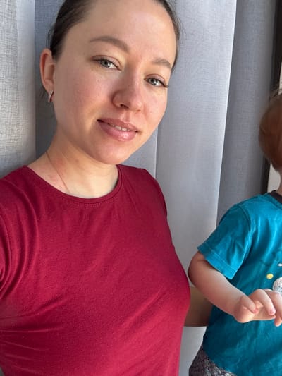 The image depicts an adult female and a young child indoors in Gamovo, Russia, positioned in front of light-colored sheer curtains. The female, centered on the left, has light skin, dark hair pulled back, light-colored eyes, and visible freckles. She wears a plain maroon short-sleeved t-shirt and a small, rectangular, possibly jewel-encrusted earring in her left ear, along with a thin necklace. Her lips are slightly parted in a faint smile, and she looks directly at the camera. To her right, a young child is partially visible from the torso up, wearing a teal short-sleeved t-shirt with a light-colored design and patterned dark shorts. The child has reddish-blonde hair and is facing generally forward. The lighting suggests natural light diffused through the curtains.