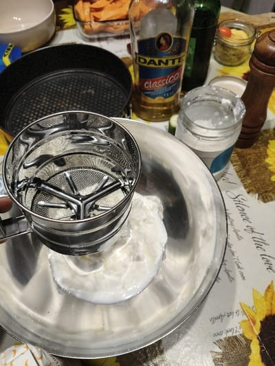 The image displays an indoor scene on a table in Villa di Gemmano, Italy, during food preparation. In the foreground, a left hand holds a stainless steel flour sifter directly above a large, reflective stainless steel mixing bowl that contains a substantial quantity of a white, creamy substance. To the left of the sifter, a dark, round baking pan with a perforated base is visible, positioned in front of a light-colored ceramic bowl. Further back on the right side of the table, a clear glass bottle of "DANTE Classico" olive oil (1L) stands next to a dark green bottle. Adjacent to these are a glass jar containing white powdered material and another small glass jar holding green herbs or spices, topped with a small red tomato. A wooden pepper mill is located on the far right. In the background, a rectangular glass baking dish filled with sliced orange root vegetables is present. The table surface is covered by a light-colored tablecloth adorned with a sunflower pattern and text, part of which reads "Silence of the love" and "to her Apollo."