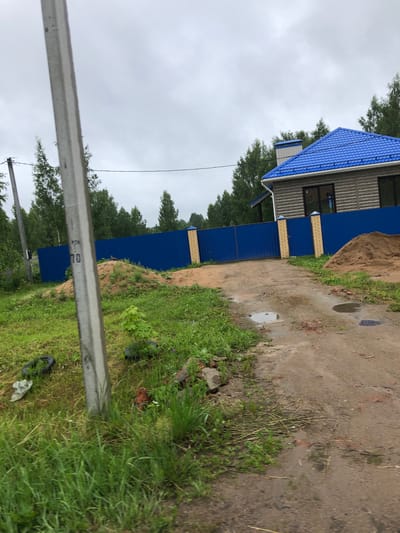 The image depicts an overcast day in an outdoor setting, likely a rural or semi-rural area near Safonovo, Russia, given the presence of a wooden-sided house with a blue metal roof and a matching blue metal fence. The property appears to be under construction or recently developed, with a large pile of sand on the right side of the dirt driveway. A concrete utility pole is prominent in the foreground on the left, with some faded markings visible on it. The ground is a mix of dirt and grass, with some puddles of water on the driveway suggesting recent rain. Green trees form a backdrop to the house and fence. There are no people or animals visible in the image. The overall impression is one of a work in progress in a somewhat undeveloped location.