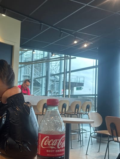 This image depicts an indoor scene within a commercial establishment, likely a cafe or restaurant, located in Saint-Louis, France.

In the foreground, the upper back and right profile of an individual wearing a black, glossy, puffer-style jacket are visible. Dark hair is pulled back, and a portion of the right ear is discernible. A clear plastic Coca-Cola bottle with a red cap, displaying "Coca-Cola GOÛT ORIGINAL" in red and white text, occupies the center foreground. The bottle is approximately two-thirds full.

The midground features a light-colored counter with multiple light wood and black metal bar stools. A silver faucet is positioned on the counter. To the right, a small round wooden table with a black base, surrounded by additional matching chairs, is visible.

Beyond the counter and tables, large windows reveal an exterior view. Outside, a complex, multi-level metal structure is present, possibly scaffolding or an architectural element. An overcast sky is visible above. Behind the counter and through the window, a male subject in a bright pink short-sleeved shirt and glasses is observed. He appears to be attending to an object on the counter. A green rectangular sign with white text, possibly an exit indicator, is visible outdoors through the window, pointing right.

The interior ceiling is dark, featuring a grid pattern and track lighting with multiple circular light fixtures.