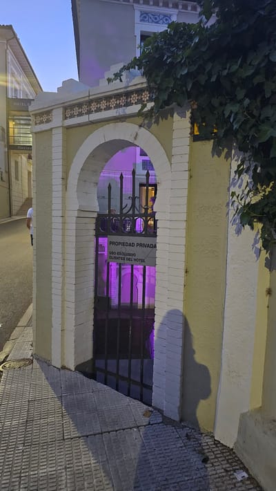 The image captures an arched entryway to a private property, illuminated by purple lighting emanating from within. A sign affixed to the black metal gate reads "PROPIEDAD PRIVADA USO EXCLUSIVO CLIENTES DEL HOTEL," indicating it is private property for the exclusive use of hotel guests. The entryway is constructed with white brick and features decorative tiling along the top. To the left, a street with tiled sidewalks is visible, with a person in a white shirt walking away from the camera. In the background, a building with the partial sign "HOTEL BA" suggests the location is a hotel. The scene appears to be in the city of Alhama De Aragón, Spain, as indicated by the provided context. The time of day is dusk, with the sky displaying a pale blue hue.