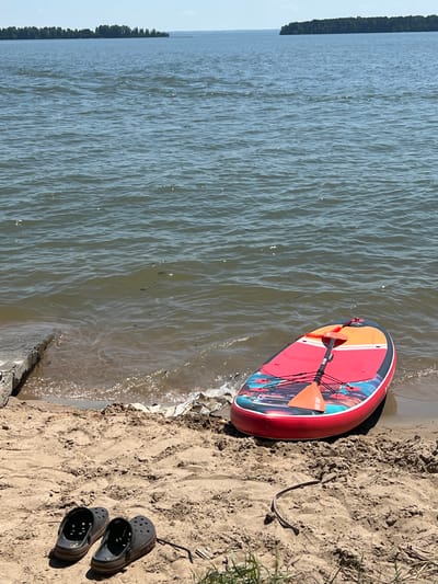 An inflatable stand-up paddleboard with an orange paddle lies partially submerged at the edge of a calm, wide body of water. The paddleboard is red with a colorful graphic design on its lower half. Dark gray clogs are placed on the sandy shore in the foreground. The background consists of a vast expanse of water under a clear sky, with a distant tree-lined shore visible on the horizon. The environment appears to be a natural lake or large river.