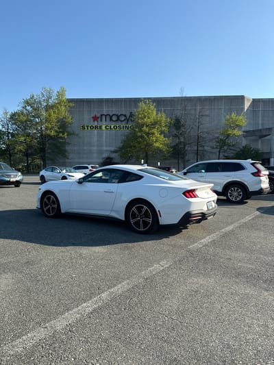 This outdoor image captures a parking lot scene in Glen Burnie, United States of America, on a bright, sunny day. The sky is clear and blue, and the strong shadows cast by vehicles suggest a mid-morning or late afternoon time.
In the foreground, a row of cars is parked on a grey asphalt lot with visible white parking lines. The most prominent vehicle is a white, two-door sports coupe, resembling a Ford Mustang, positioned closest to the viewer. Its rear and side are visible, featuring dark multi-spoke wheels and distinct taillights. Behind it, a white SUV, possibly a Honda CR-V, is also parked. A Maryland license plate is visible on the sports coupe. Further down the parking row, other vehicles are partially visible, including a dark-colored SUV or sedan.
The backdrop is dominated by a large, multi-story concrete building, characteristic of a department store. A prominent sign on the building clearly reads "Macy's" in red and black lettering with its iconic star logo. Directly below, in large yellow capital letters with a black outline, the text "STORE CLOSING" is displayed. Several trees with green foliage and some bare branches are scattered around the base of the building and between the parked cars. No people are visible in the frame.
The overall scene depicts a typical shopping center environment, with the "Macy's STORE CLOSING" sign being a significant focal point, indicating a specific event for the retail location.