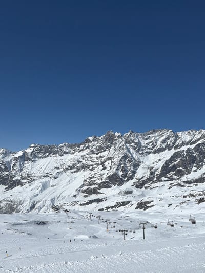 The scene depicts a snow-covered alpine ski resort in Valtournenche, Italy, under a clear, deep blue sky. A large, rugged mountain range, heavily covered in snow and ice with exposed dark rock formations, dominates the background and upper mid-ground. In the mid-ground and foreground, a wide, groomed ski slope is visible, showing tracks from previous activity. Multiple ski lift lines ascend the slope, featuring dark grey support poles and empty chair mechanisms. Several small, dark figures, identified as skiers or snowboarders, are scattered across the lower portion of the ski slope, engaged in descent or standing. The environment is bright, indicating direct sunlight and clear weather conditions.