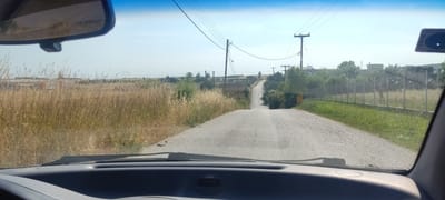 The image presents a driver's perspective from inside a car, looking forward onto a rural or semi-rural road in Λάκκωμα, Greece. The immediate foreground shows the car's dashboard, windshield wipers, and the bottom edge of the rearview mirror. The sky is bright and clear, indicating daytime, likely sunny, with some distant haze.

The cracked, paved road stretches directly ahead, gently sloping uphill and disappearing over a crest in the distance. On both sides of the road, particularly on the left, are extensive fields of dry, tall, golden-brown grass. Some patches of greener, shorter vegetation are visible closer to the road, especially on the right.

Overhead, a network of electrical power lines stretches across the sky, supported by several wooden utility poles lining the road. On the right side, a chain-link fence runs parallel to the road, separating it from an area with more green grass and some sparse trees. Further down on the right, a large, dark sign with light-colored Greek text appears, seemingly reading "NEA DIASTASIS" (New Dimension). A smaller yellow sign is also visible but its text is illegible.

In the midground, the landscape includes scattered bushes and trees. The background features several indistinct buildings, possibly industrial or agricultural structures, partially obscured by trees and atmospheric haze. The overall impression is a quiet, sun-drenched, somewhat arid landscape. A sticker with unreadable text is affixed to the top right of the windshield. No people are visible, and no specific activity beyond driving or being present in the car is occurring.