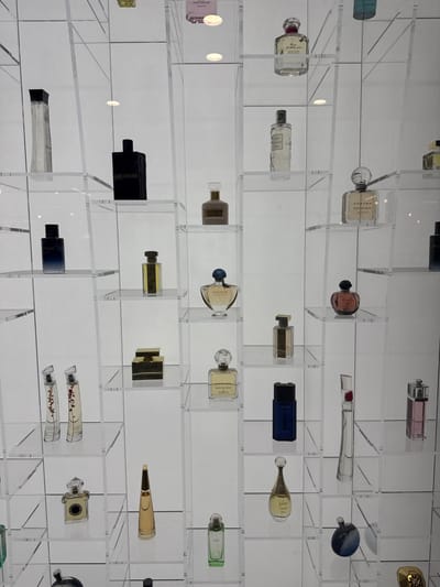 The image displays a brightly lit, transparent acrylic or glass shelving unit showcasing approximately 30-40 perfume bottles against a white background. The display is organized in a grid pattern, with each individual compartment holding one to two distinct bottles.
**Objects:**
The perfume bottles vary significantly in shape, size, and color. Identifiable brands include:
* A light pink, square Marc Jacobs bottle in the top left.
* A black, rectangular bottle labeled "RAD & VOILAR" on the second row, second from the left.
* A Guerlain bottle with a distinctive clear, textured, round cap on the second row, second from the right.
* A dark blue rectangular bottle, possibly Sauvage, on the third row, first from the left.
* A gold-colored Dolce & Gabbana box set on the fourth row, center-left.
* A Carven bottle, specifically "DANS MA BULLE," with a spherical cap on the fourth row, second from the right.
* A J'adore Dior bottle, recognizable by its elongated, golden-capped, tear-drop shape, on the sixth row, second from the right.
Liquids within the bottles range from colorless to various shades of amber, yellow, blue, and pink. Bottle materials are predominantly clear glass, with some opaque black or dark blue designs.
**Environment:**
The scene is an indoor commercial or private display, characterized by clean lines and clear materials. The white background is uniformly illuminated, suggesting either backlighting or integrated lighting within the display structure. There are no individuals, actions, or interactions depicted.
**Location:**
The scene is located within the city of Saint-Gilles - Sint-Gillis, Belgium.