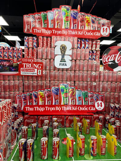 The image shows a retail display of Coca-Cola cans, featuring designs related to the FIFA World Cup and national teams. The display is predominantly red and white, with various cans stacked in pyramid formations. Some cans are adorned with national flags and football-themed graphics. A prominent red banner at the top reads "Thu Thập Trọn Bộ Thiết Kế Quốc Gia" (Collect the Full Set of National Designs). Below this, another red banner states "CƠ HỘI TRÚNG* 9 Bộ Gaming Lenovo Legion" (Chance to Win* 9 Lenovo Legion Gaming Sets). The background shows more stacked Coca-Cola products and what appears to be a portion of a large Coca-Cola logo resembling a soccer ball. The display is set up on a green surface patterned like a soccer field. A small number "14 LỌN" (14 Bottles) is visible in a red circle on the right side of the display. The context of Phu Thuy, Vietnam, is not directly discernible from the image itself, but it is a detail provided as a location context for the scene. There are no people visible in the image.