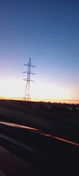 An electrical tower stands prominently against a gradient sky, transitioning from deep blue to a fiery orange. The tower's intricate lattice structure is sharply silhouetted. A strip of land, possibly a field or forest, stretches horizontally along the base, darkening towards the right. Hints of a road or pathway are visible, framed by the edge of a vehicle window, creating a sense of movement and perspective. The sunset casts a warm glow, highlighting the landscape's textures.