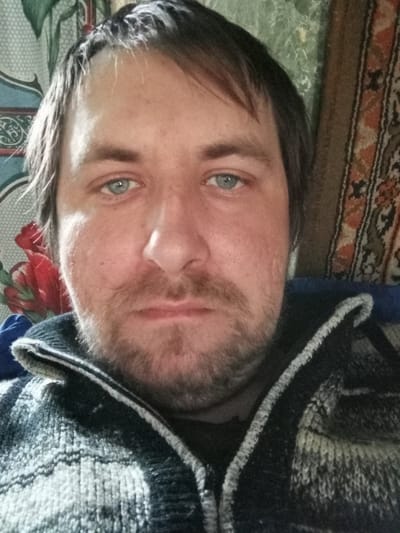 A self-portrait of a Caucasian male with fair skin, blue eyes, and dark brown hair that is partially receding at the hairline. He has a full, untrimmed beard and mustache. He is wearing a knitted sweater with a zipper, likely in a casual setting. The background is cluttered with floral-patterned and woven tapestry. There is no information in the image to determine the location is Parabel, Russia.