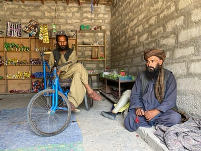 Two men are seated in a small shop in Panjwai, Afghanistan. The shop is constructed of brick and has a wooden shelf unit that is stocked with various goods. One man is seated on a blue, three-wheeled bicycle, while the other man is seated on the ground, his right leg missing from the knee down and replaced by a prosthetic. They both appear to be looking away from the camera. The shop is located at latitude 31.579 and longitude 65.568. The floor of the shop is concrete and there is a blue tarp on the ground in front of the men.