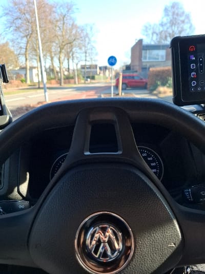 The image presents a street view in Goirle, Netherlands, from the driver's perspective inside a Volkswagen vehicle. The central foreground is occupied by a black steering wheel featuring a chrome Volkswagen emblem. Behind the steering wheel, a speedometer dial is partially visible, displaying numerical markings up to 180 km/h. To the far right, a black electronic device, likely a telematics unit or tablet, is mounted and displays various illuminated icons, including a prominent red warning symbol.

Through the windshield, a paved road and an adjacent red-colored bicycle path are visible under direct sunlight. A blue circular road sign with a white bicycle pictogram, indicating a mandatory bicycle lane, is mounted on a pole to the right of the path. A red sport utility vehicle is parked or stopped on the right side of the street, partially obscured by a low hedge. The background includes residential buildings and numerous bare deciduous trees, with a clear blue sky overhead. No individuals are visible.