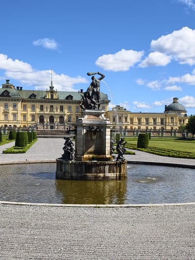 The image shows the fountain in front of Drottningholm Palace, located in Ekerö kommun, Sweden. The fountain, made of bronze, is the central subject, with water spraying upwards and outwards. Several sculpted figures are visible on the fountain, including a larger central figure holding a spear. Smaller figures are positioned around the base of the fountain, some appearing to interact with water. The fountain is situated in a circular pond, surrounded by a gravel pathway. In the background, the pale yellow facade of Drottningholm Palace is visible. The palace has a symmetrical design with multiple windows and a prominent clock tower in the center. To the left and right of the palace, manicured gardens with rows of conical hedges and flat green lawns are present. The sky is a bright blue with scattered white clouds, indicating a sunny day. The overall scene depicts a formal palace garden with a significant water feature.