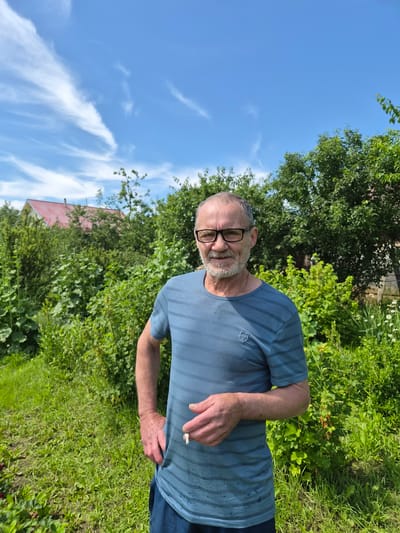 An older man with a gray beard and glasses is standing in a garden. He is wearing a blue striped t-shirt and dark blue shorts. He is holding a cigarette in his right hand. The background is a lush green garden with trees and a house with a red roof. The sky is blue with some white clouds. The image appears to be taken in the city of Novosisoevka, Russia, during the daytime.