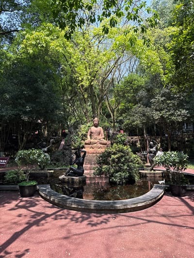 The image captures a serene scene within a park in Shizhong, China. A large, tan-colored statue of Buddha sits prominently in the middle ground, partially obscured by the leaves of a large tree. Adjacent to the Buddha statue, there's a semi-circular pond with statues of figures. The pond is edged with a dark gray trim. The right side of the image shows a reddish-brown paved pathway, covered with shadows. The environment is lush with greenery. The image is rotated 90 degrees clockwise.