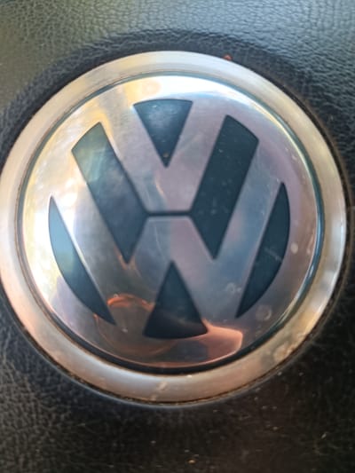 The image shows a close-up of the Volkswagen logo, which is a metallic emblem set into a black, textured surface, likely part of a car's interior such as a steering wheel. The logo consists of a stylized "VW" within a circle. The metallic surface of the logo is reflective, showing some light and color distortions, indicating it is likely polished chrome or a similar material. The black background has a fine, grained texture, typical of leather or a synthetic material used in automotive interiors. The lighting appears to be ambient, possibly from an overhead light source or daylight, creating subtle reflections and shadows on the logo. There is no text visible in the image. The scene is static and focused on the automotive emblem, suggesting a product shot or a detail captured for identification or aesthetic purposes. The location context of Nordkirchen, Germany, suggests the image was taken there, but this cannot be inferred from the visual content alone.