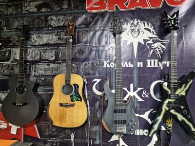 Four guitars are mounted on a wall covered with a dark, textured, brick-like fabric backdrop. The backdrop also features a large, abstract, purple and white graphic print. On the far left is a black acoustic guitar. Next to it is a natural wood acoustic guitar. To the right of that is a dark gray electric bass guitar. On the far right is a black electric guitar with a pointed, angular body, decorated with a lightning bolt design. The scene appears to be indoors. There is no visible indication of the city of Pogranichny, Russia within the image itself.