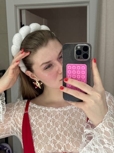 A woman holds a smartphone with a pink "pop-it" on the back, obscuring her face slightly. She wears a white lace top with a red undergarment, and a white fluffy headband. A large, star-shaped earring is visible on her left ear. The woman has fair skin and red nail polish. The background includes a window and a hint of a mirror selfie setting.