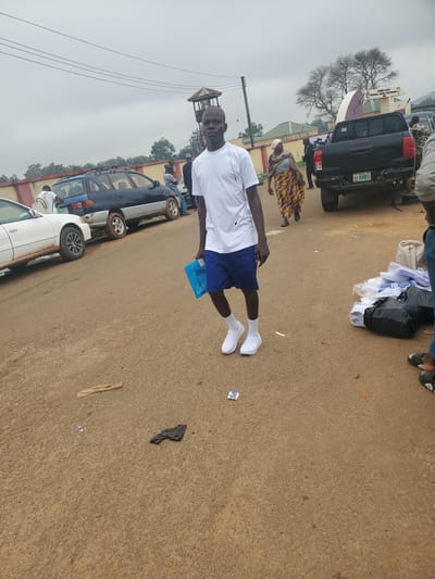 A young Black male wearing a white t-shirt, blue shorts, white socks, and white sneakers walks on a dirt road, holding a blue bag in his right hand. He is in Ohit, Nigeria. In the background, several parked cars and a dark-colored pickup truck are visible. A woman in a patterned dress and headscarf walks in the background to the left of the pickup truck. A guard tower and a building with text that reads "JESUS GATE MAXWELL HOUSE COUNTRY CLUB" are also in the background. The ground is covered in dirt and small debris, including a plastic bag and a crumpled piece of paper. A pile of white clothing, possibly uniforms, is stacked on the ground to the right of the pickup truck.