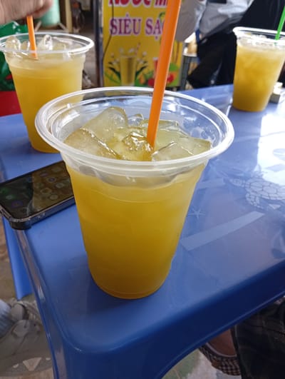 The image shows a blue plastic table in Phú Hòa, Vietnam, holding three clear plastic cups filled with a yellow liquid, possibly sugarcane juice, and ice. Each cup has a straw. An iPhone rests on the table next to the central cup. In the background, a menu sign with yellow and orange text is visible, along with indistinct figures of people. A portion of someone's clothing is seen on the lower right side of the image. The surface of the table has a faint pattern visible beneath the cups.