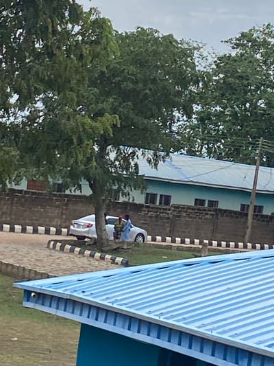The image depicts an outdoor scene in Sabo, Nigeria.  A large green tree dominates the left side of the frame. Adjacent to the tree, a building with a light green exterior and a corrugated roof is visible. A white car parked in front of the building appears to have belongings loaded on top of it. A person dressed in blue is next to the car. A black and white striped border runs parallel to the building. On the right, the corner of another building with a blue roof is visible. The sky is overcast.