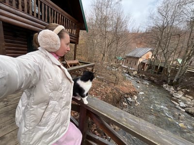 The scene is situated in Crkvine, Montenegro, portraying a female individual and a bicolored cat on a wooden balcony overlooking a river. The woman, positioned on the left, is seen in profile, wearing a light-colored (likely beige or off-white) puffer jacket, a pink garment underneath, and large, fluffy beige earmuffs. Her left arm is extended forward, holding a mobile device to capture a self-portrait. A black and white cat stands on the weathered wooden railing directly in front of the woman, oriented towards the distant landscape. The cat features a white chest, belly, and paws, with black fur on its back, head, and tail. Behind the woman, a log cabin structure is partially visible. Below the balcony, a moderately wide river flows rapidly over a rocky bed. On the opposite bank, a stone-faced building with a sloped, dark gray metallic roof stands amidst deciduous trees, which are largely bare of foliage, indicating a late autumn or winter season. Several smaller structures with green roofs are visible further along the riverbank. The background consists of densely wooded hills under an overcast sky.