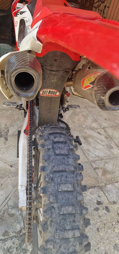 The image displays a detailed, rear-facing perspective of a red and white dirt bike or motocross motorcycle, described as being located in Kolarovo, Bulgaria. The motorcycle is stationary, parked outdoors on a paved surface.
The main subject is a used, off-road motorcycle. Its rear bodywork and seat are predominantly red. White plastic side panels are visible, with one on the left featuring a black "CRF" logo and appearing to have a repair with white tape. The exhaust system consists of dual silencers; the left one has a carbon fiber end cap, and the right one also features a carbon fiber end cap and a prominent "PRO CIRCUIT" sticker in yellow, red, and black on its metallic body. A black, gritty plastic under-fender is present above the rear wheel, bearing an oval "THE ORIGINAL HOT RODS CRANKS AND RODS" sticker in red and white. Two metallic, serrated footpegs are visible on either side of the bike. The drivetrain includes a metal chain, partially visible on the left side, appearing dirty. The rear wheel is equipped with a heavily treaded, knobby off-road tire that shows wear and dirt accumulation.
The motorcycle is situated on an outdoor ground surface composed of light-colored, possibly concrete or stone, tiles arranged in a geometric pattern. The background is out of focus but appears to be an indistinct structure or wall. The scene is illuminated by natural light, indicative of daytime. No individuals are present in the image, and no actions are depicted.