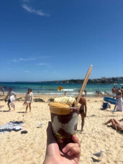 A person holds a dessert cup containing banana slices, a waffle, and a dark sauce, topped with a wooden spoon. The background shows a sunny beach scene with sand, the ocean, and people enjoying themselves. The sky is a clear, bright blue.