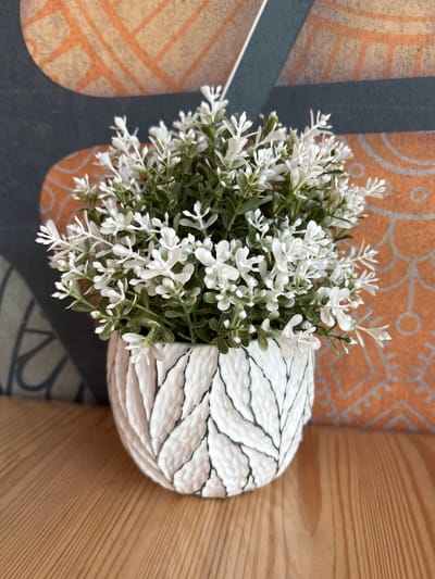 An artificial white flowering plant in a textured, white pot with black leaf-like patterns sits on a light brown wooden surface. The pot is round and bulbous. Behind the pot is a wall with a patterned wallpaper featuring orange and dark gray geometric shapes. The image is a close-up shot, focusing on the plant and pot, with the background slightly out of focus. The scene appears to be indoors, possibly in a home or office setting. The city of Inzer, Russia, is not visible or inferable from the image.