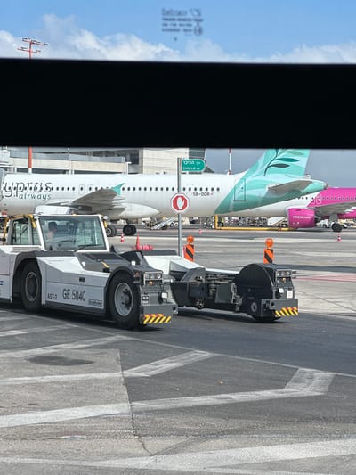 An airport tarmac scene is depicted under a clear blue sky with scattered white clouds. In the foreground, a white and gray tow tractor, labeled "AST-2 GE 5040 Goldhofer," is positioned on the asphalt. The tractor has a driver in the cab wearing an orange vest. Behind the tractor, part of a black towing attachment is visible.
In the middle ground, a Cyprus Airways airplane with a distinctive teal leaf logo on its tail is parked, connected to a jet bridge. To the right of this plane, a Wizz Air airplane with a pink fuselage is visible. Various airport equipment, including cones and a yellow hose, are scattered on the tarmac. A green directional sign reads "דרך הכיכר CIRCLE RD."
The background shows airport structures and more aircraft. A black rectangular object, possibly a shadow or a device obstructing the view, is present in the upper portion of the image, obscuring the top of the airport buildings and the sky. The overall impression is a functional airport environment during the day.