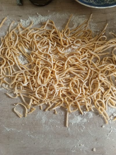 A close-up, overhead shot shows a pile of freshly made, uncooked pasta noodles scattered across a light-colored countertop dusted with flour. The noodles are golden yellow and appear to be hand-cut, varying slightly in thickness and length. They are entangled and curved, with some pieces lying flat and others looping. The flour creates a light dusting around and on top of the noodles, suggesting they have recently been prepared. The countertop has a subtle texture and appears to be made of a smooth material. In the upper right corner of the image, a portion of a blue and white patterned plate is visible, suggesting that the noodles might be prepared for a meal or to be stored. The lighting is natural and soft, casting minimal shadows. The scene is devoid of any specific elements that would indicate a location in Buraly, Russia.
