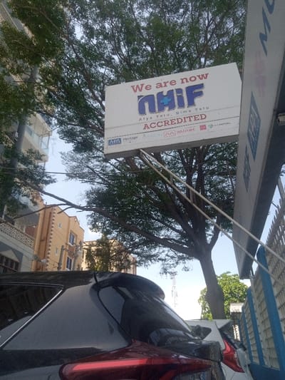 The image depicts an outdoor urban scene in Majengo ward, Kenya, viewed from a low-angle perspective looking upwards towards the sky and surrounding structures. A prominent white rectangular sign, supported by white metal struts, occupies the upper-center of the frame. The sign displays "We are now NHIF Aiya Yetu, Bima Yetu ACCREDITED" in red and blue lettering, followed by logos for APA, Heritage, LAISON, Minet, and O.MUTUAL.

A large deciduous tree with green foliage dominates the upper background, its branches extending across the clear blue sky, partially obscuring parts of a light-colored multi-story building on the far left and a light brown building with white trim further back. In the immediate foreground, the rear portion of a dark-colored (likely black or dark grey) SUV or crossover vehicle is visible, featuring a red taillight and a rear spoiler. A white vehicle is partially visible behind and to the right of the dark SUV. A vertical structure with blue accents is partially visible on the far right, and a lattice communication tower is faintly discernible in the distant background. No individuals are present in the depicted scene.