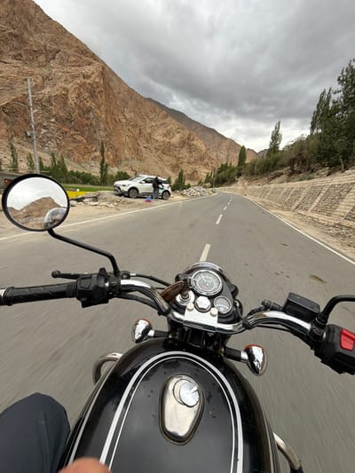 A first-person perspective shot from a motorcycle shows a person riding on a road. The rider's hands are on the handlebars, and the front of the motorcycle, including the fuel tank and instrument cluster, is visible. The road curves to the right, flanked by a dry, rocky mountainside on the left and trees and a stone-built retaining wall on the right. In the middle distance, a white SUV is parked on the side of the road, with two people standing near it. The sky is overcast with gray clouds. The scene appears to be a rural or mountainous area, possibly Likche, India, given the dry climate and mountainous terrain.