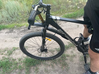 This is a medium shot, eye-level photograph of the front section of a black bicycle. A person's legs and lower torso are visible on the right side of the frame, suggesting they are either about to ride or have just dismounted. The bicycle has a black frame, black handlebars with textured grips, and a front fender. The front wheel, with its knobby tire, is prominent in the lower left of the image. The bicycle appears to be on a dirt path with sparse green vegetation and dry grass on either side. The lighting suggests it is daytime, possibly late afternoon or early evening, with soft, natural light. The overall scene is casual and outdoors, possibly in a rural or semi-rural area like Заречный ауылдық округі, Kazakhstan.
Visible text includes the brand name "GRAND" on the fork of the bicycle and a partially visible "adidas" logo on the person's shorts. There are also reflective elements on the bicycle, one of which is orange. The scene conveys a sense of simple outdoor activity.