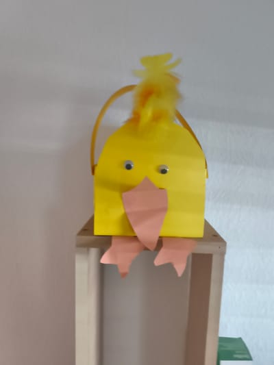 This is a media file of a handmade yellow chick craft, likely made for Easter. The chick is bright yellow and has googly eyes, a pink paper beak, and pink paper feet. A yellow feather tuft adorns the top of its head. A yellow ribbon handle is attached to the top of the chick's body. The chick is sitting on a light brown wooden shelf. In the background, a white wall is visible. The lighting suggests it is daytime, and the setting appears to be indoors. There is no text visible in the image. The overall impression is a cute and cheerful craft, representative of springtime or Easter decorations.