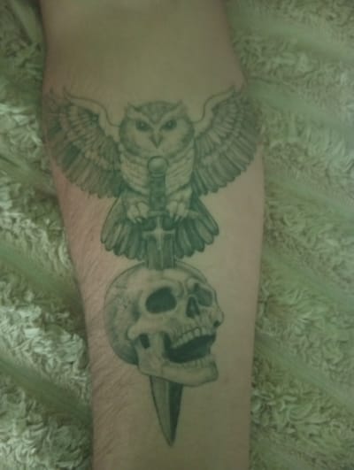The image shows a close-up of a tattoo on a person's arm. The tattoo depicts an owl with outstretched wings perched above a skull with an open mouth. A dagger is positioned vertically between the owl and the skull, with its hilt above the owl's talons and its blade piercing through the skull. The tattoo is rendered in grayscale. The background of the image is a fuzzy, light-colored fabric, possibly a blanket or upholstery. The setting is indoor, and the time of day and weather are not discernible. No text is visible in the image.