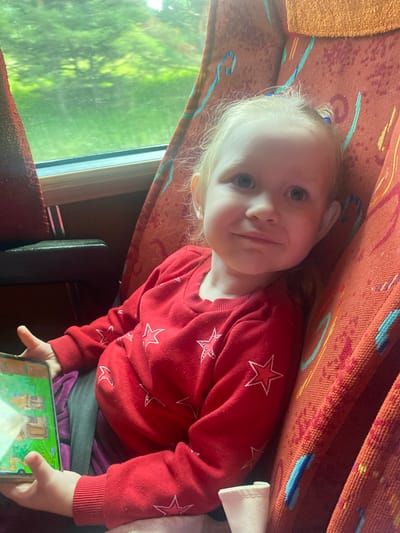The image shows a young girl, likely a toddler, sitting in a bus seat. She is wearing a red sweater with white stars and has her hair tied in two small ponytails. The girl is looking towards the camera with a slight smile and is holding a mobile device in her left hand. The bus interior is visible, with orange and red patterned seats and a window showing greenery outside. The setting is a bus, and no specific details suggest it is in Osinovka (Polozovskii S/S), Russia.