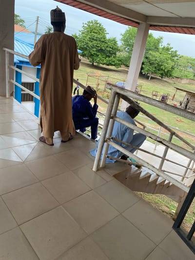 The image shows three men on a building's tiled balcony or landing. Two men are seated on the steps leading down, behind a rusty metal railing. One man is wearing a light blue robe and the other a darker blue robe and a hat. A third man, wearing a brown robe and a hat, stands on the tiled floor, facing away from the camera. Outside the railing, there is a grassy area with trees and what appear to be metal frames of some kind. A blue and white building is visible in the lower left. The overall scene suggests an outdoor, possibly public, space. The location of Sabo, Nigeria is mentioned.
