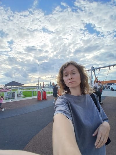 A medium shot, selfie-style photograph captures a woman with shoulder-length, wavy brown hair from the chest up. She is wearing a grey t-shirt and stands in an outdoor area with a paved ground. In the background, a playground is visible with swings and other equipment. Several people are present, including children on the swings and adults standing nearby. A covered structure with a dark roof is also in the background, along with lampposts and a distant road with cars. The sky above is filled with dramatic clouds against a blue sky. The scene appears to be a public park or recreational area.