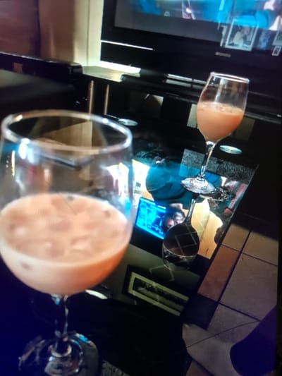 The scene portrays an indoor, residential environment in Soweto, South Africa. In the foreground, a clear stemmed glass holds a light orange, opaque liquid with multiple ice cubes. A second, identical stemmed glass containing a similar beverage with ice is positioned on a dark, highly reflective surface, likely a coffee table, in the midground. This table reflects a television screen displaying predominantly blue content, a person wearing a blue top, and a metallic object with a crosshatch pattern. The television set, framed in black, is located in the background. The floor in the lower right consists of square, light-colored tiles, and a human foot, appearing to wear a white sock, is partially visible on it. No direct human figures are clearly visible; human presence is indicated by reflections and a visible foot.