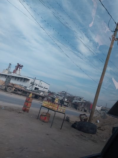 An oblique wide shot captures a street scene in a-Zawayda, Palestinian Territory, under a sky with scattered cirrus clouds and multiple overhead utility lines. The viewpoint is from inside a vehicle, indicated by reflections on a window pane in the upper right and a dark dashboard/door frame in the lower right.

In the foreground, a dusty ground surface is visible. A makeshift roadside stall features a metal table displaying approximately two dozen plastic bottles containing a yellow liquid, likely cooking oil, arranged in rows. A brown cardboard box rests beneath the table. To the left of the table, two stacks of red and brown cardboard boxes stand vertically. To the right, a large deflated tire or similar dark, rounded object lies on the ground next to a wooden utility pole equipped with insulators and wires.

The midground features an asphalt road running horizontally across the frame. On the opposite side of the road, a large white industrial building with a flat roof and red structural elements, possibly machinery or vents, is prominent. Arabic text on the building reads "مصنع عثمانية للأعلاف" (Othmanieh Feed Mill/Factory). Several large trucks, including flatbed types, are parked or in motion along the road and adjacent to the building. One dark-clad human figure is discernible near the trucks on the right side of the road. Piles of debris, possibly scrap metal, are visible behind the vehicles and building. The background is indistinct, showing a hazy horizon.