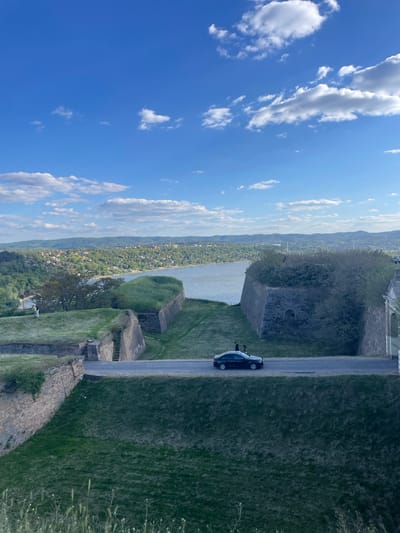 This high-angle, wide shot captures a panoramic view from Petrovaradin Fortress in Serbia, looking over the Danube River and the city of Novi Sad. The image is taken on a clear, sunny day with a bright blue sky dotted with scattered white cumulus clouds.
In the foreground, on the right side of the frame, are parts of the ancient stone fortifications of the fortress, including moss-covered walls and grassy mounds. A narrow, paved road or path runs horizontally across this area. A dark-colored car, possibly a sedan, is parked or slowly moving on this path. Two individuals are standing near the car; one is clearly visible next to the driver's side, appearing to lean on the vehicle or look inside.
The midground features the wide, calm expanse of the Danube River, its surface reflecting the sky. Across the river, the urban landscape of Novi Sad stretches out, characterized by numerous buildings nestled amidst dense greenery. Beyond the city, a long range of hills or mountains, also covered in trees, extends into the hazy distance, marking the horizon.
The overall scene suggests a leisurely visit to a historical landmark, with the people seemingly taking a break by their car to enjoy the expansive vista. No visible text is present in the image.